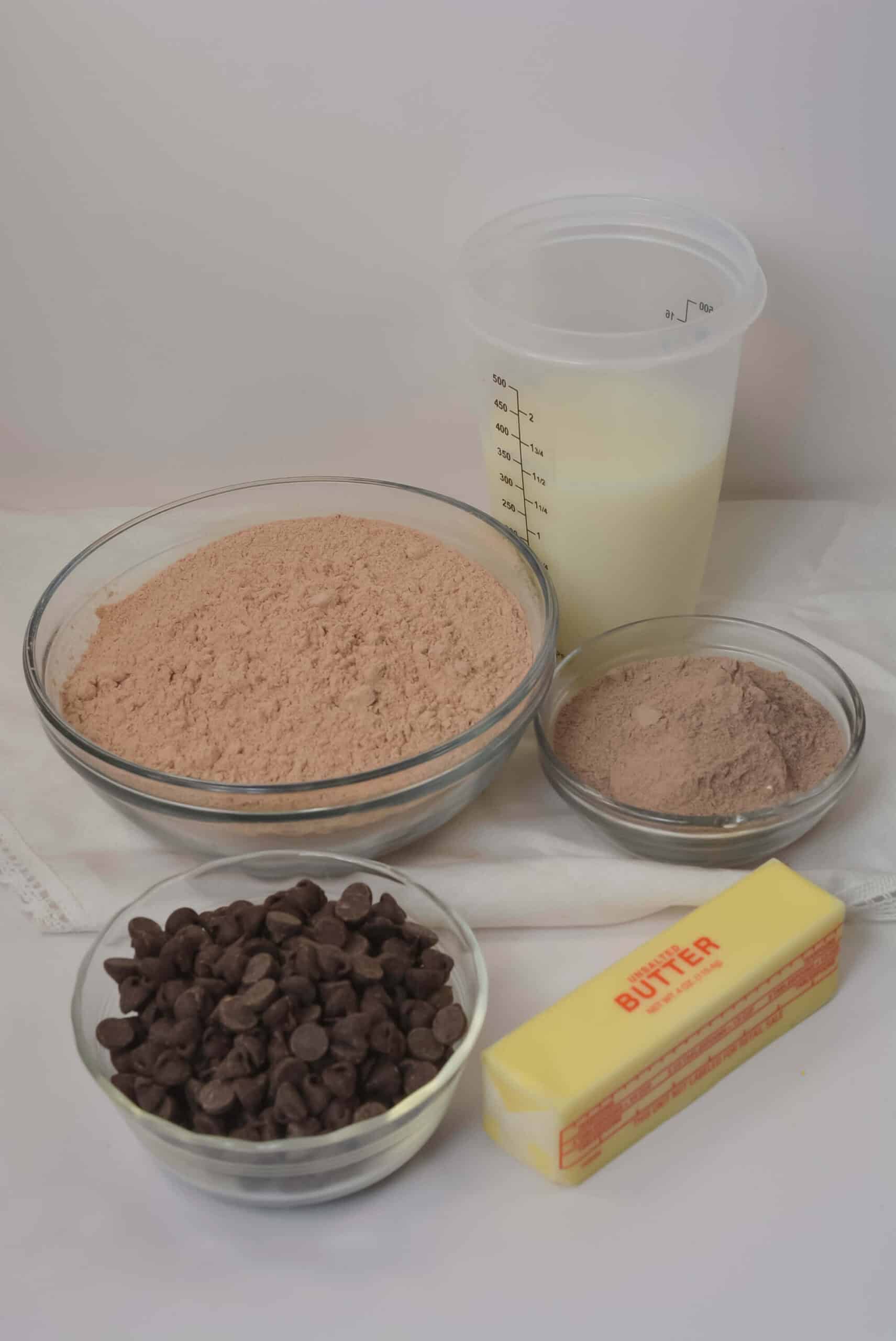 Five baking ingredients on a white surface: a bowl of  mix,  pudding mix, chocolate chips, a stick of butter, and milk—everything you need for the dessert.