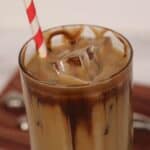 A glass of caramel iced coffee with milk and chocolate drizzle, filled with ice cubes, and topped off with a red-and-white striped straw.