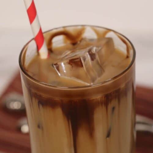 A glass of caramel iced coffee with milk and chocolate drizzle, filled with ice cubes, and topped off with a red-and-white striped straw.