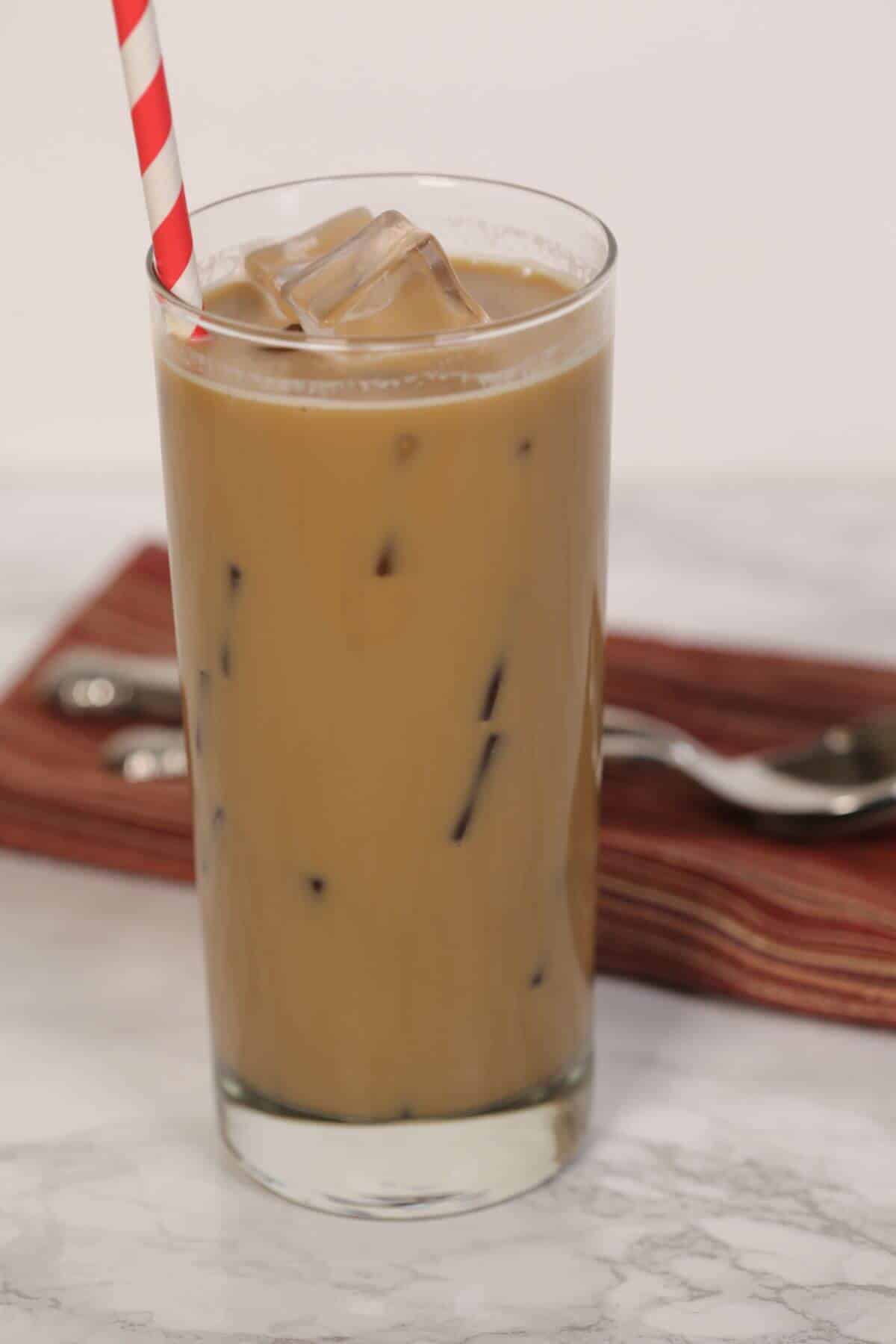 A tall glass of caramel iced coffee with a striped straw sits on a marble surface, accompanied by a folded napkin and spoon.