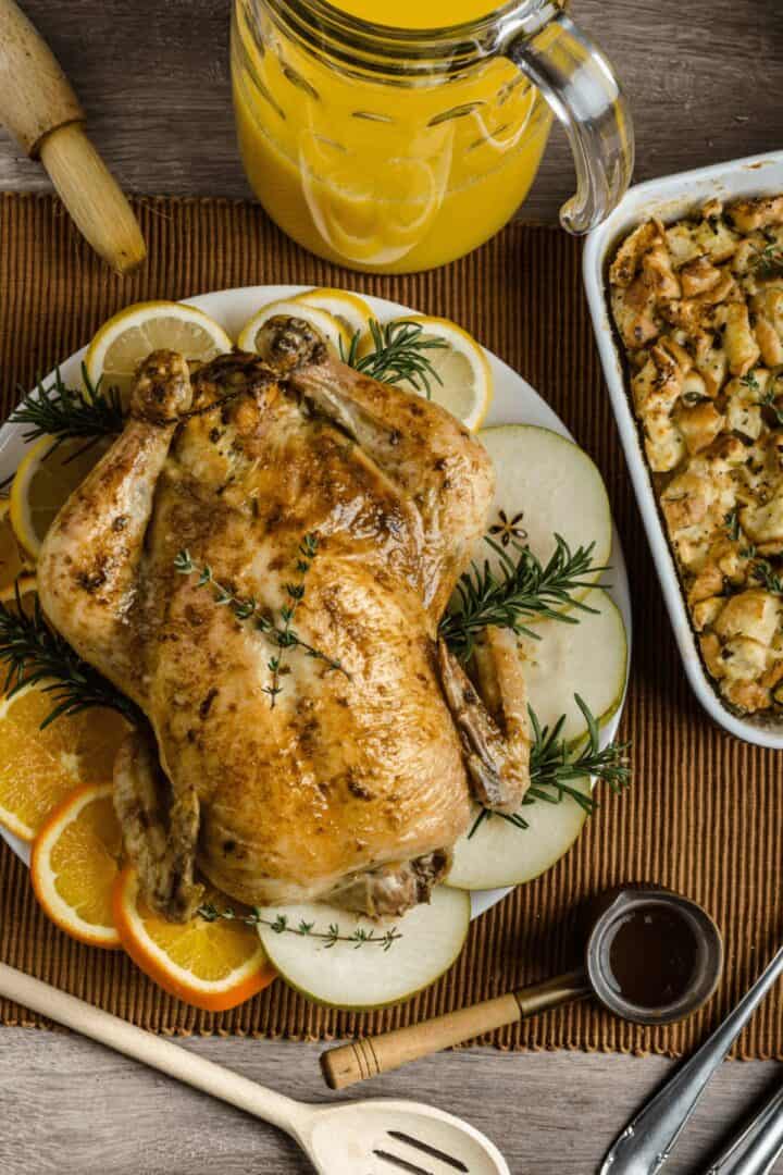 Roast chicken garnished with orange, apple, and rosemary on a plate, next to a baking dish of stuffing. A pitcher of orange juice and wooden utensils rest on a brown placemat—perfect with Citrus Turkey Brine flavors.
