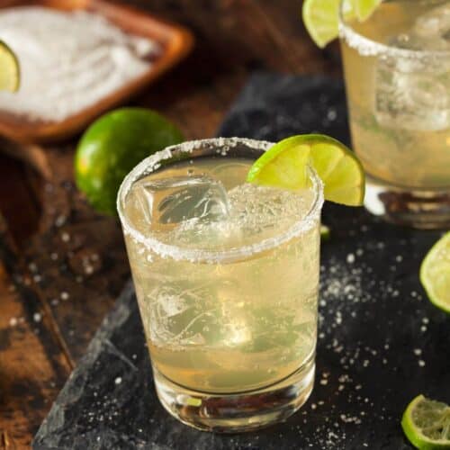 A glass with a salted rim, ice cubes, and a lime wedge garnish sits on a dark surface, surrounded by scattered lime slices and salt.