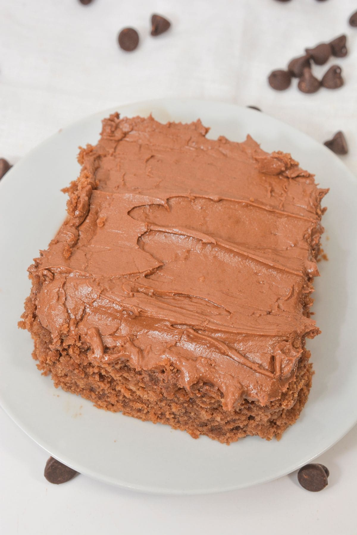 A rectangular piece of Cocoa Mayonnaise Cake with chocolate frosting sits on a white plate, with chocolate chips scattered in the background.