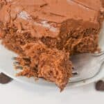 A close-up of a fork holding a bite of Cocoa Mayonnaise Cake with chocolate frosting, with the rest of the cake on a white plate in the background.