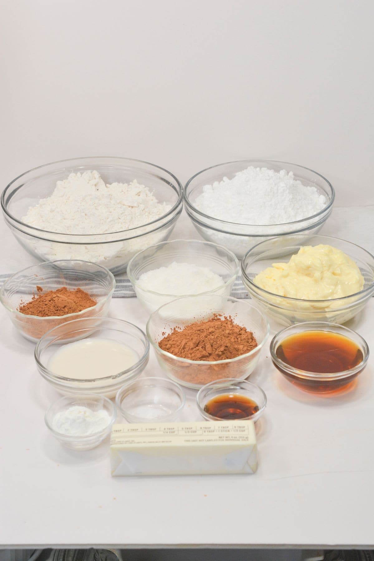 Various baking ingredients including flour, powdered sugar, butter, cocoa powder, milk, vanilla extract, and baking powder, are arranged in clear glass bowls on a white surface.