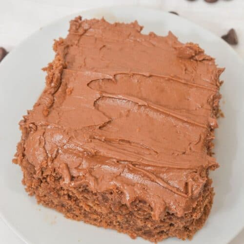 A rectangular piece of Cocoa Mayonnaise Cake with chocolate frosting sits on a white plate, with chocolate chips scattered around.