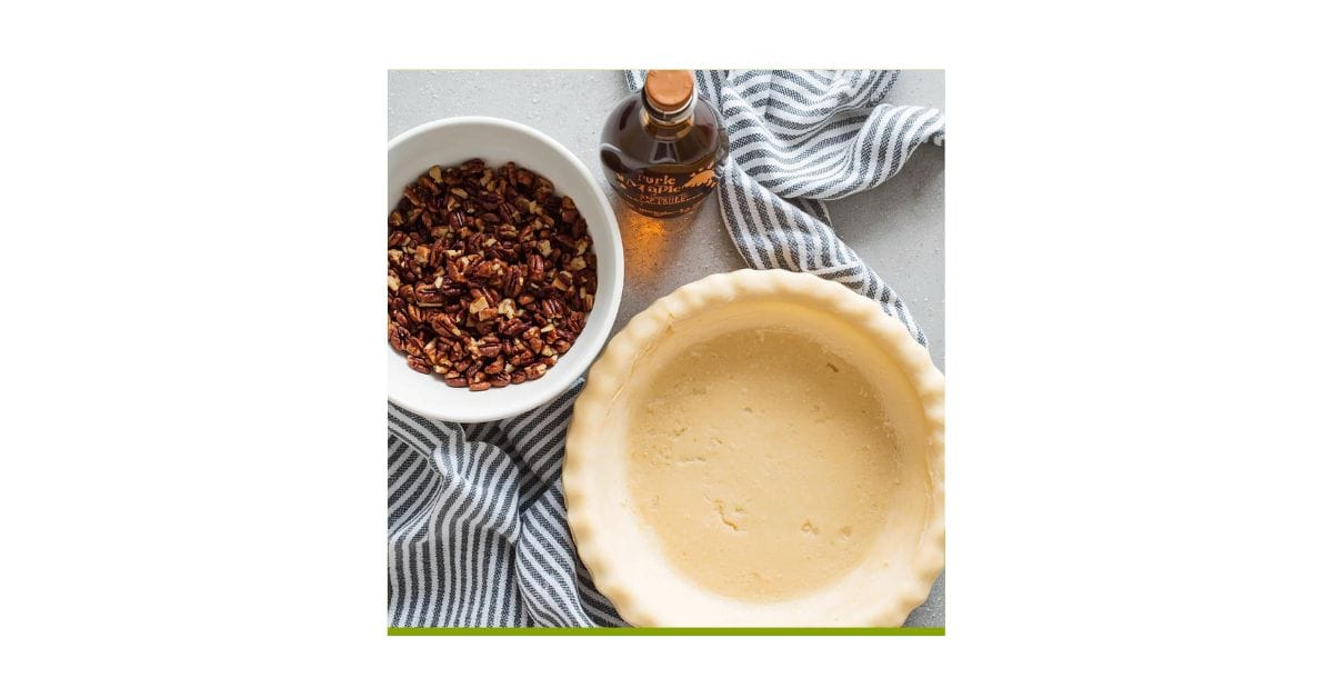 A bowl of chopped pecans, a bottle of maple syrup, and an unbaked pie crust await on a striped cloth—everything you need to create the perfect Maple Syrup Pecan Pie.
