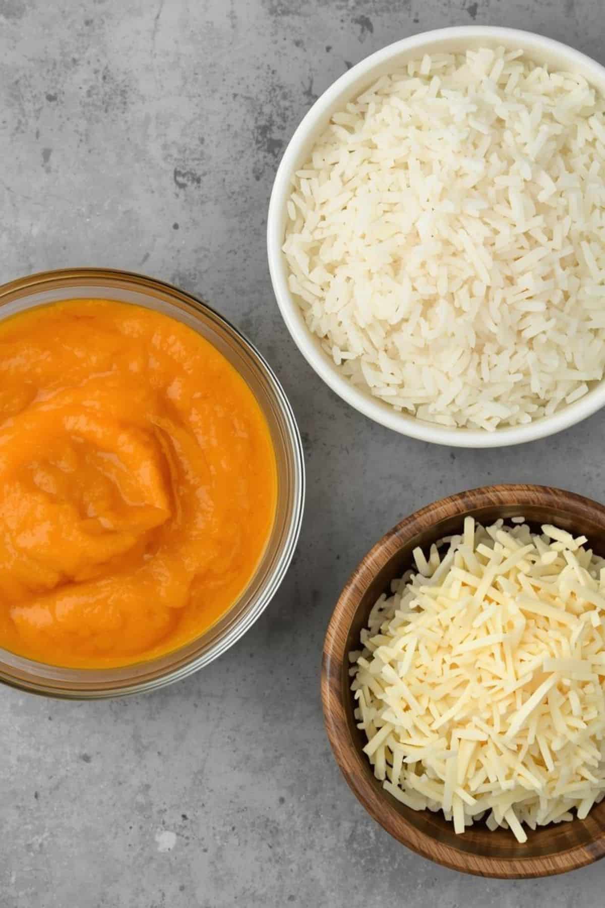 Three bowls on a gray surface: one with cooked white rice, one with shredded cheese, and one with orange pumpkin puree..