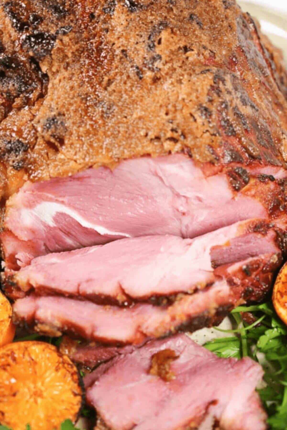 Close-up of a copycat honey baked ham with several slices cut, showcasing its juicy pink interior. Grilled orange halves and greens are visible in the foreground.