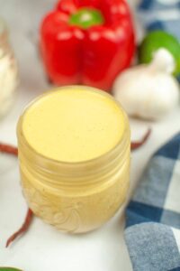 A small glass jar filled with creamy yellow sits on a white surface, with a red bell pepper, garlic, and a blue plaid cloth nearby.