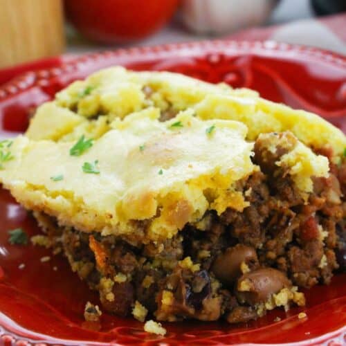 A serving of cornbread-topped chili pie casserole with ground meat and vegetables on a red plate, garnished with chopped herbs.