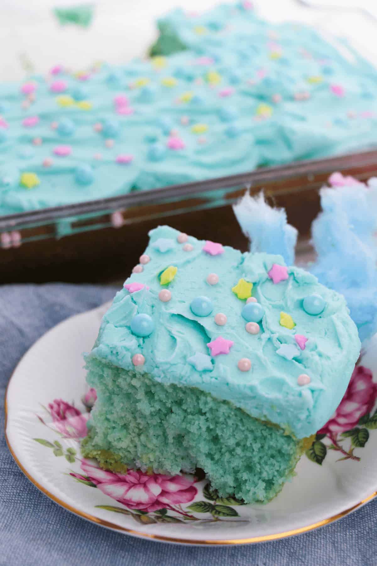 A slice of blue-frosted cotton candy cake with star and pearl sprinkles rests on a floral plate, while a tray of similarly delightful cakes lingers in the background.