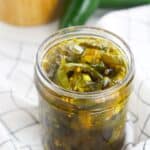 A glass jar filled with sliced Cowboy Candy pickled jalapeños in liquid, placed on a white cloth with a black grid pattern.