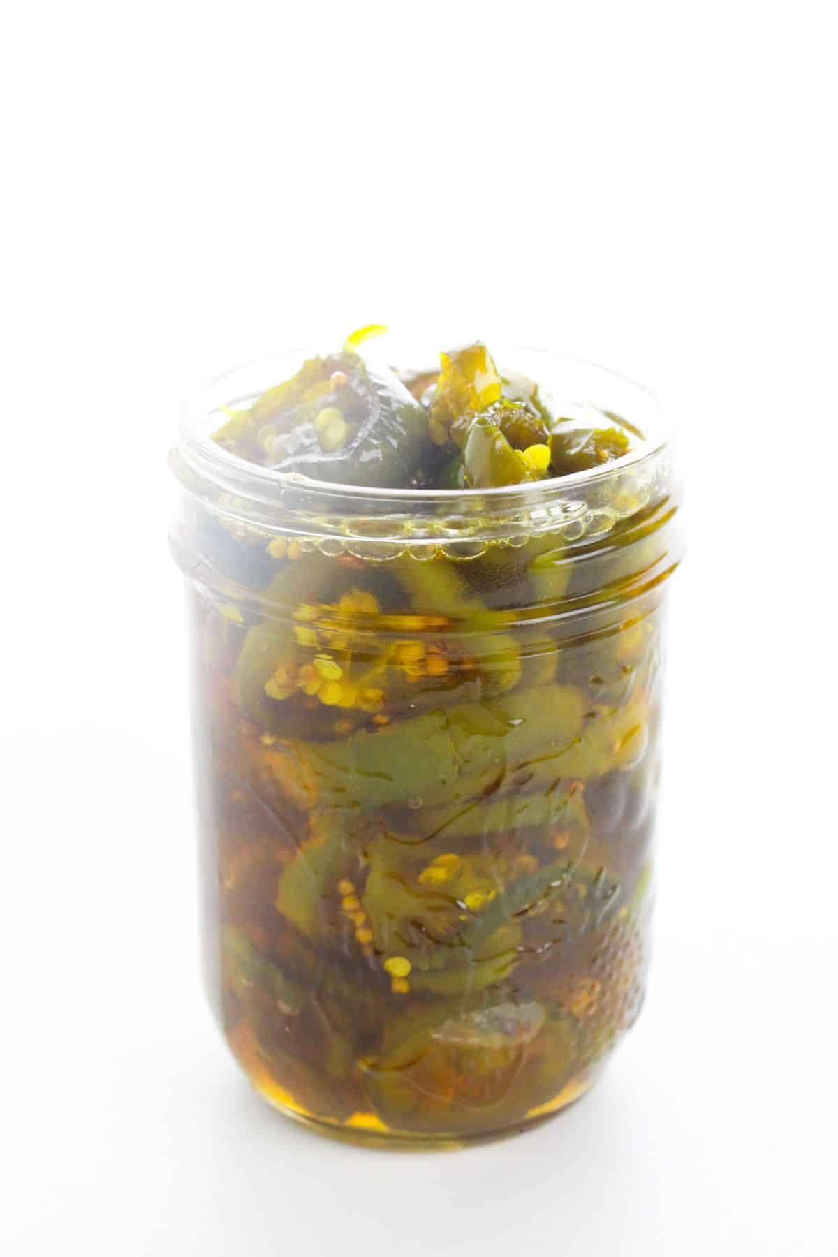 A glass jar filled with sliced  jalapeños in brine, placed against a white background.