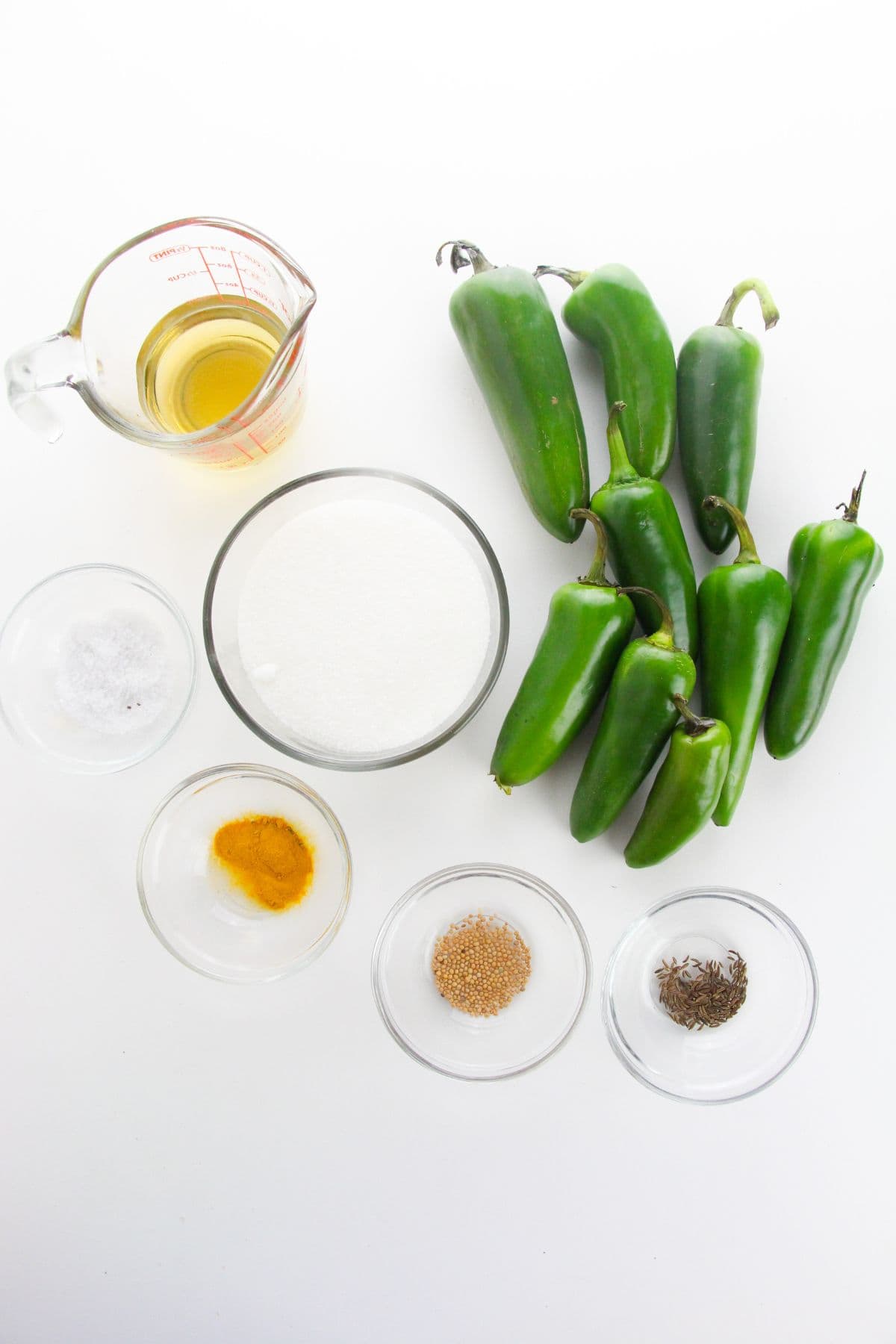A group of fresh jalapeños, a measuring cup of vinegar, a bowl of sugar, salt, turmeric, mustard seeds, and cumin seeds are arranged on a white surface.