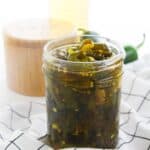 A glass jar filled with Cowboy Candy pickled jalapeños sits on a white checkered cloth, with a salt container and a bottle in the background.