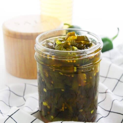 A glass jar filled with Cowboy Candy pickled jalapeños sits on a white checkered cloth, with a salt container and a bottle in the background.