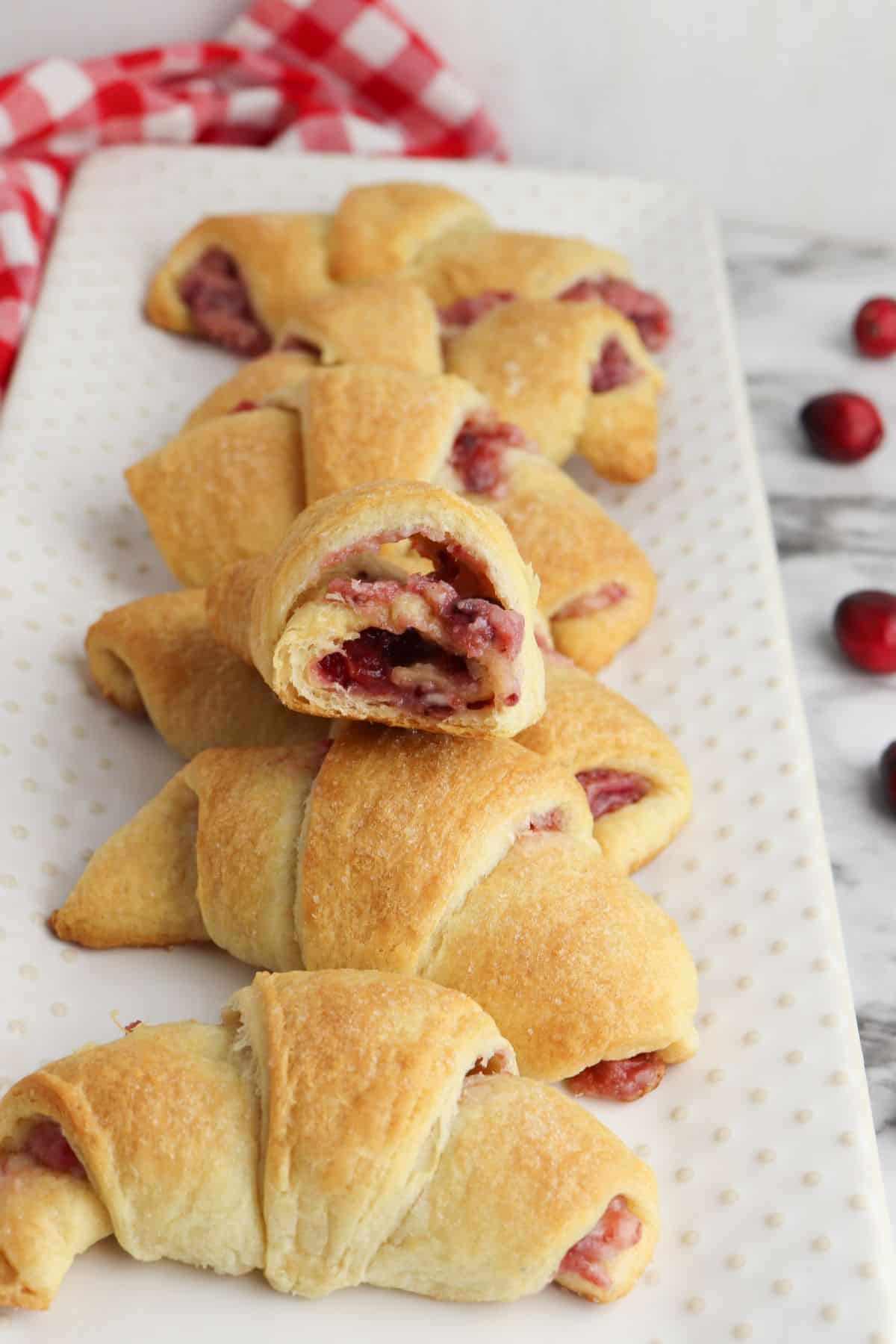 A row of golden-brown Cheesecake Crescent Rolls filled with cranberry filling is arranged on a white rectangular tray, with one roll split open to reveal its creamy, tangy inside.