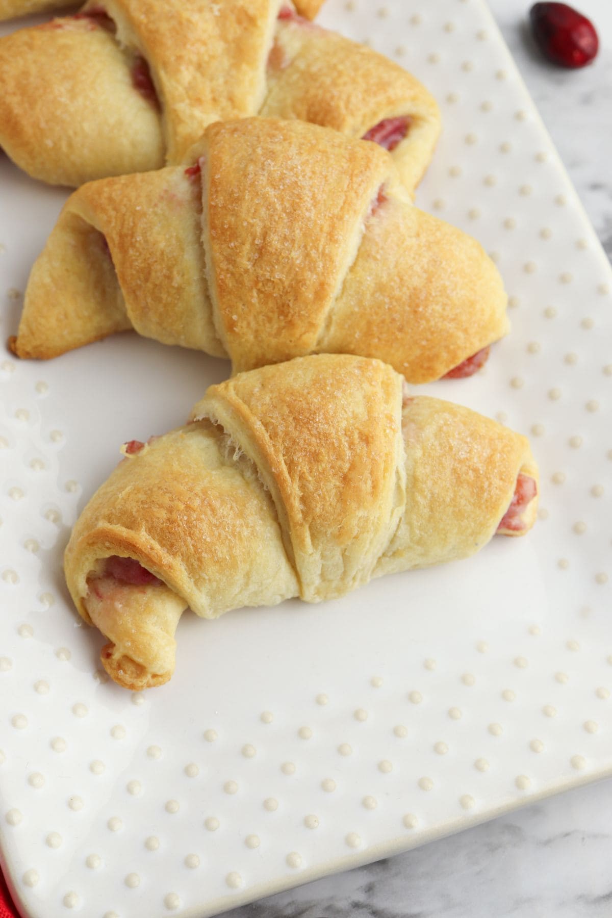 Three golden-brown Cheesecake Crescent Rolls with a visible fruity filling are arranged on a white, textured rectangular plate.