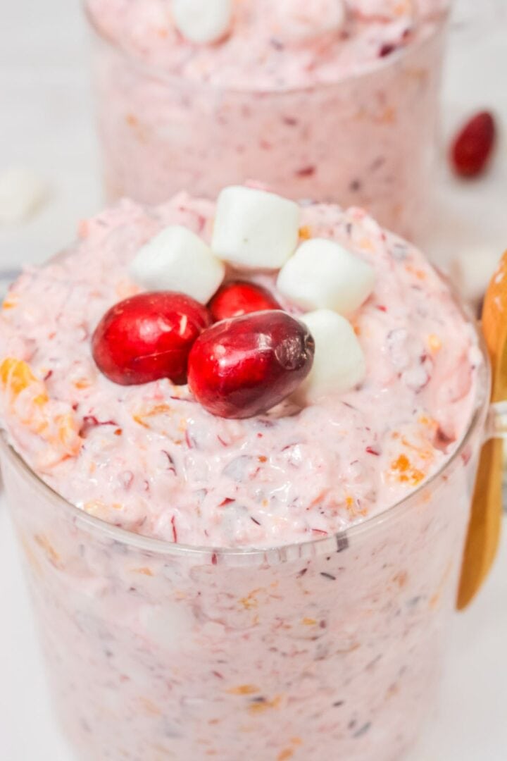 A glass filled with a pink creamy Cranberry Fluff salad mixture, topped with three cranberries and three mini marshmallows—an irresistible holiday dessert.