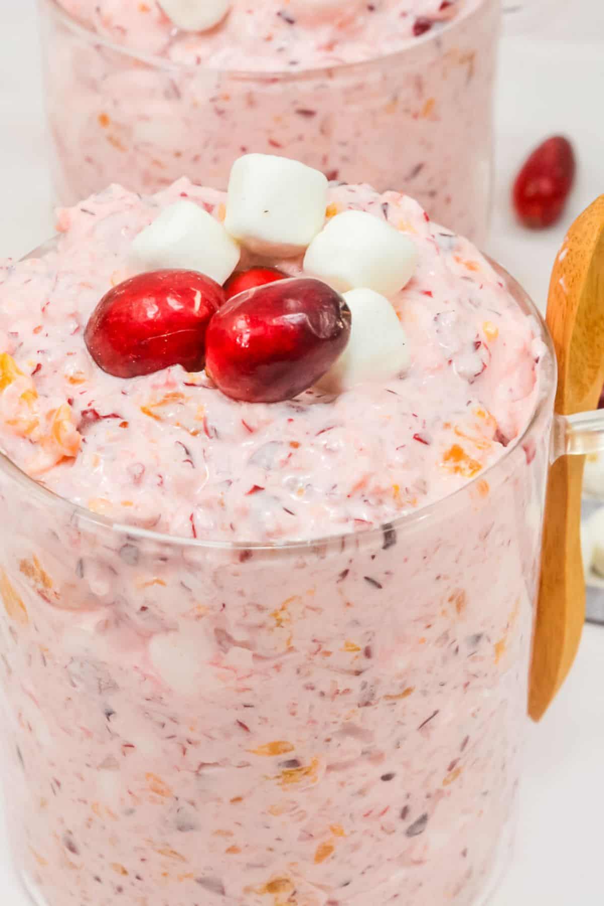 Glass cup filled with pink Cranberry Fluff dessert, topped with two cranberries and three mini marshmallows, with a wooden spoon and some ingredients in the background—perfect as a festive holiday recipe.