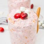 A glass cup filled with pink Cranberry Fluff, topped with cranberries and mini marshmallows, rests beside a wooden spoon. Another cup of this festive holiday dessert and scattered garnish appear in the background.