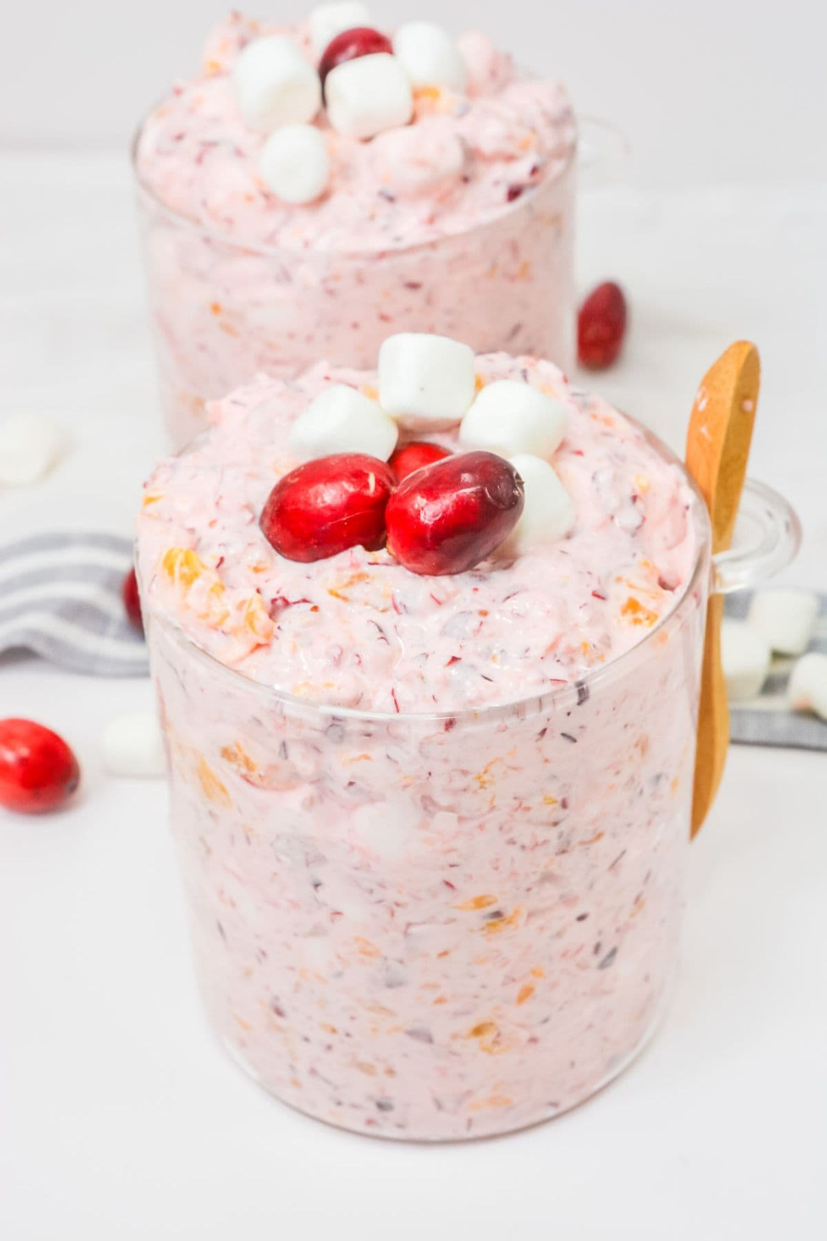 A glass cup filled with pink Cranberry Fluff, topped with cranberries and mini marshmallows, rests beside a wooden spoon. Another cup of this festive holiday dessert and scattered garnish appear in the background.