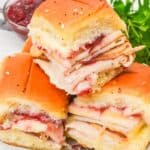 Three Cranberry Turkey Sliders with cheese and cranberry sauce are stacked on a white plate, garnished with parsley, with a bowl of cranberry sauce in the background.