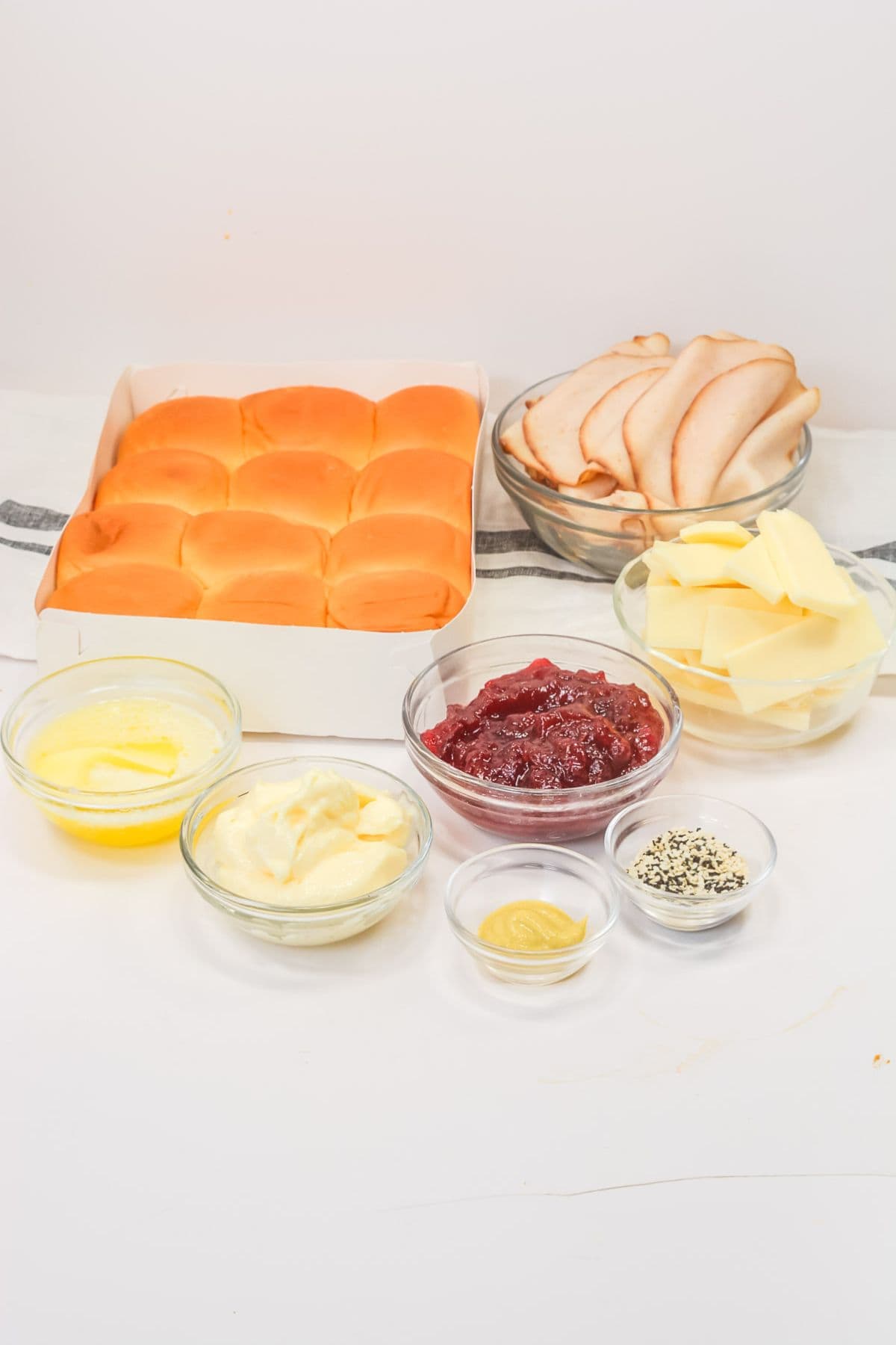 Ingredients for making Cranberry Turkey Sliders arranged on a white surface, including slider buns, sliced turkey, cheese, butter, mayonnaise, cranberry sauce, mustard, and seasoning.