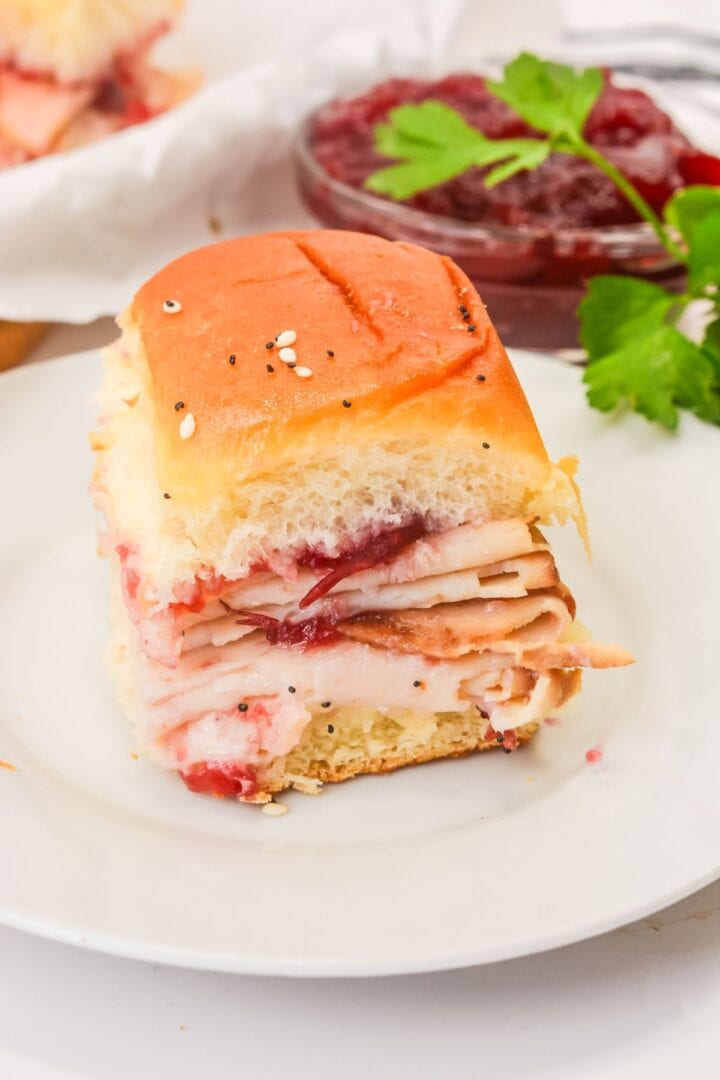 A Cranberry Turkey Slider with poppy seeds on a soft roll, served on a white plate with cranberry sauce and parsley in the background—perfect for those who love classic Cranberry Turkey Sliders.