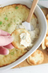 A hand holds a cracker with Cream Cheese and Crab Dip, poised above a white dish brimming with the creamy baked delight. Chopped green onions adorn the top, while a wooden spoon peeks from the side.