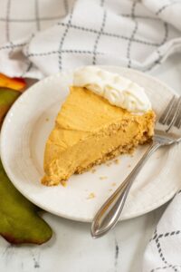 A slice of Cream Cheese Pumpkin Pie cheesecake with a graham cracker crust and whipped cream on top, served on a white plate with a fork.