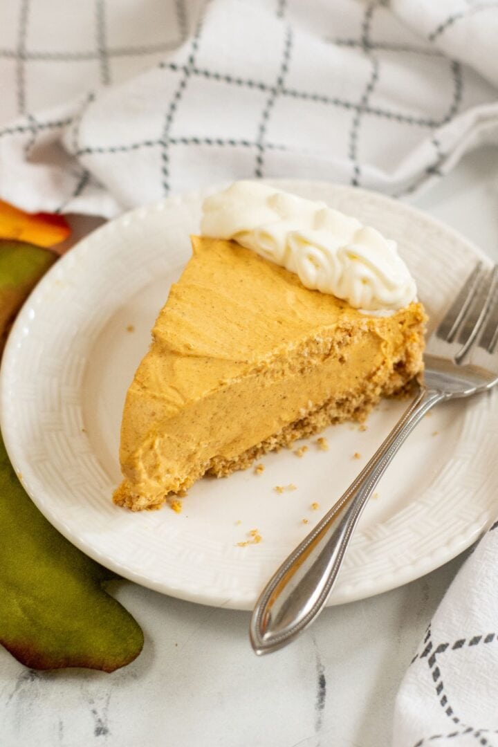 A slice of Cream Cheese Pumpkin Pie cheesecake with a graham cracker crust and whipped cream on top, served on a white plate with a fork.