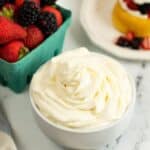 A bowl of Cream Cheese Whipped Cream sits on a marble surface next to a carton of fresh strawberries and blackberries and a plate with a dessert topped with fruit and cream.