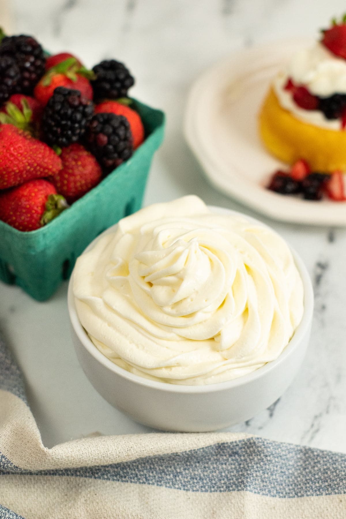 A bowl of Cream Cheese Whipped Cream sits on a marble surface next to a carton of fresh strawberries and blackberries and a plate with a dessert topped with fruit and cream.