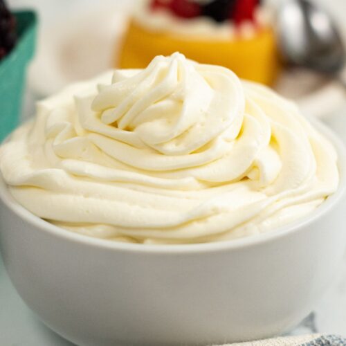 A white bowl filled with swirled Cream Cheese Whipped Cream sits on a table, with desserts and a spoon blurred in the background.