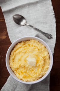 A bowl of creamy mashed potatoes with a hint of parmesan, topped with a pat of butter on a cloth napkin. A spoon is placed beside the bowl.