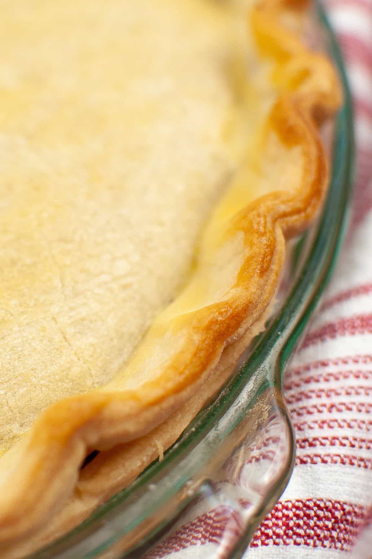 A close-up of a baked pie with a golden-brown, crimped Crisco Pie Crust in a glass dish, resting on a red and white striped cloth.