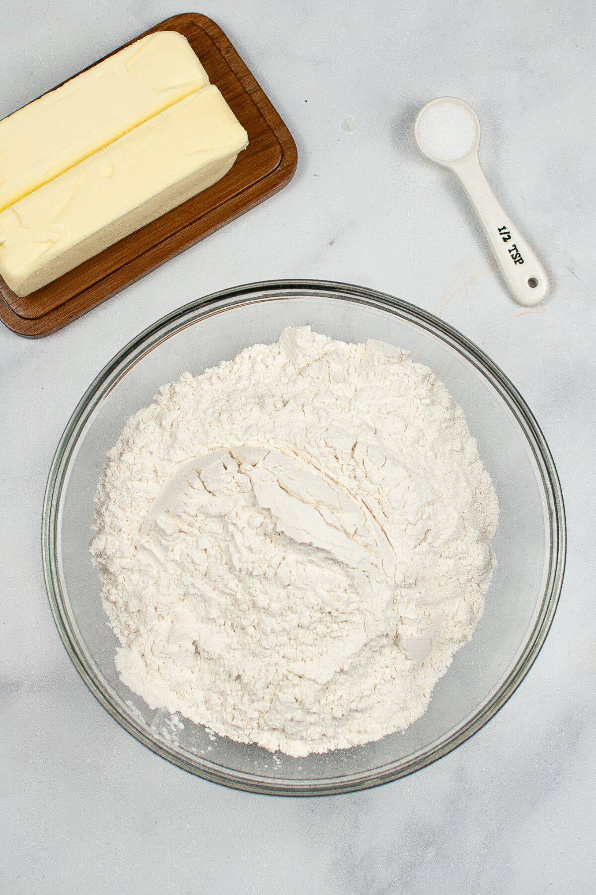 A glass bowl filled with flour, a wooden tray with two sticks of butter, and a tablespoon of salt on a white surface.
