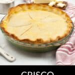 A baked pie with a golden-brown Crisco Pie Crust sits in a glass dish on the counter, surrounded by butter, flour, and a striped towel. Text reads "Crisco Pie Crust.