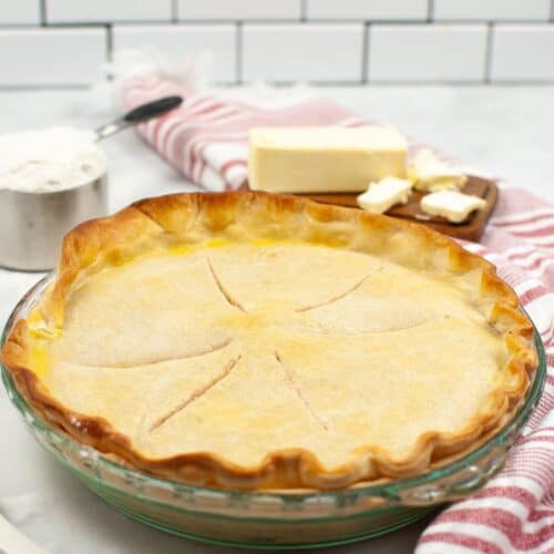 A baked pie with a golden Crisco Pie Crust in a glass dish, with butter, flour, and a striped towel in the background on a white kitchen counter.