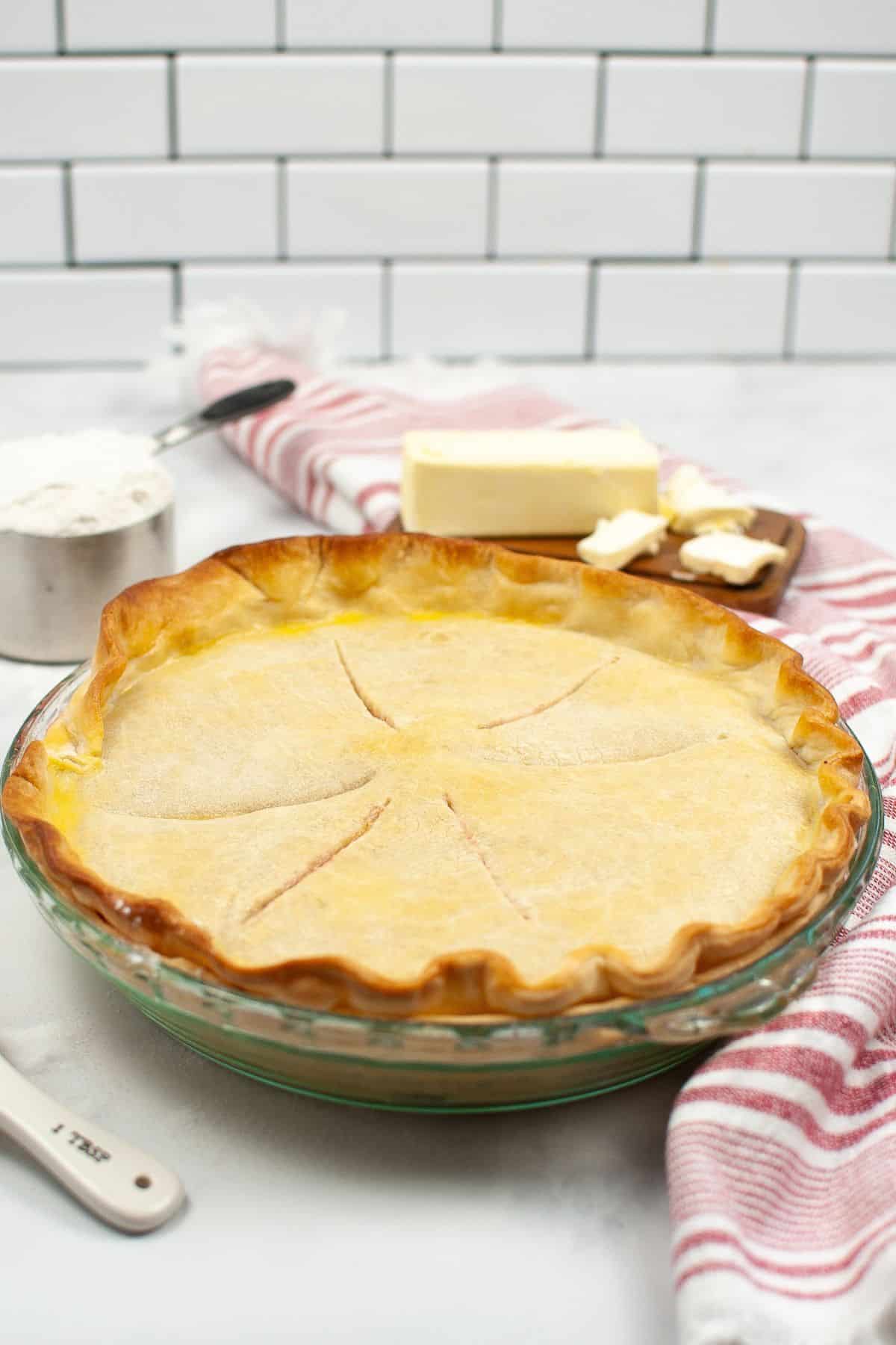 A baked pie with a golden Crisco Pie Crust in a glass dish, with butter, flour, and a striped towel in the background on a white kitchen counter.