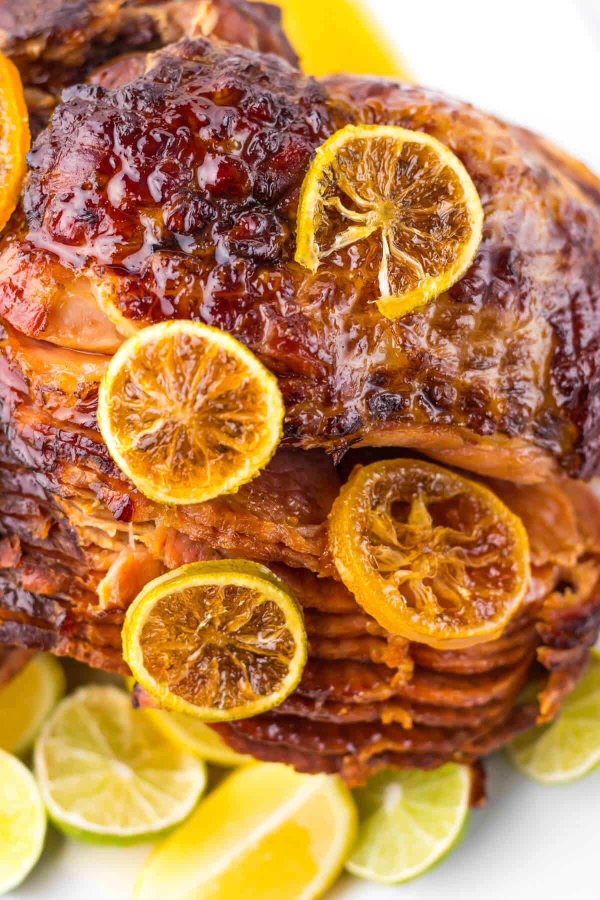 Crock Pot Brown Sugar Pineapple Ham shines with its sweet glaze, topped with caramelized lemon slices and surrounded by fresh lemon and lime wedges on a plate.