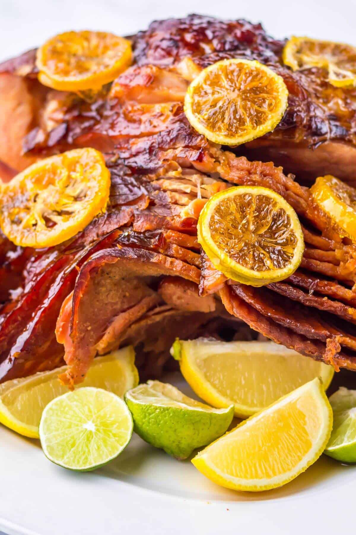 A delightful Crock Pot Brown Sugar Pineapple Ham, beautifully sliced and glazed, topped with lemon slices and encircled by lime and lemon wedges on a pristine white plate.