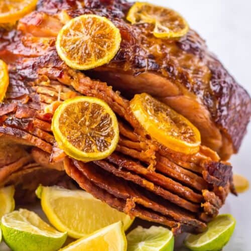 A Crock Pot Brown Sugar Pineapple Ham, adorned with orange slices and encircled by lime wedges, sits enticingly on a white plate.