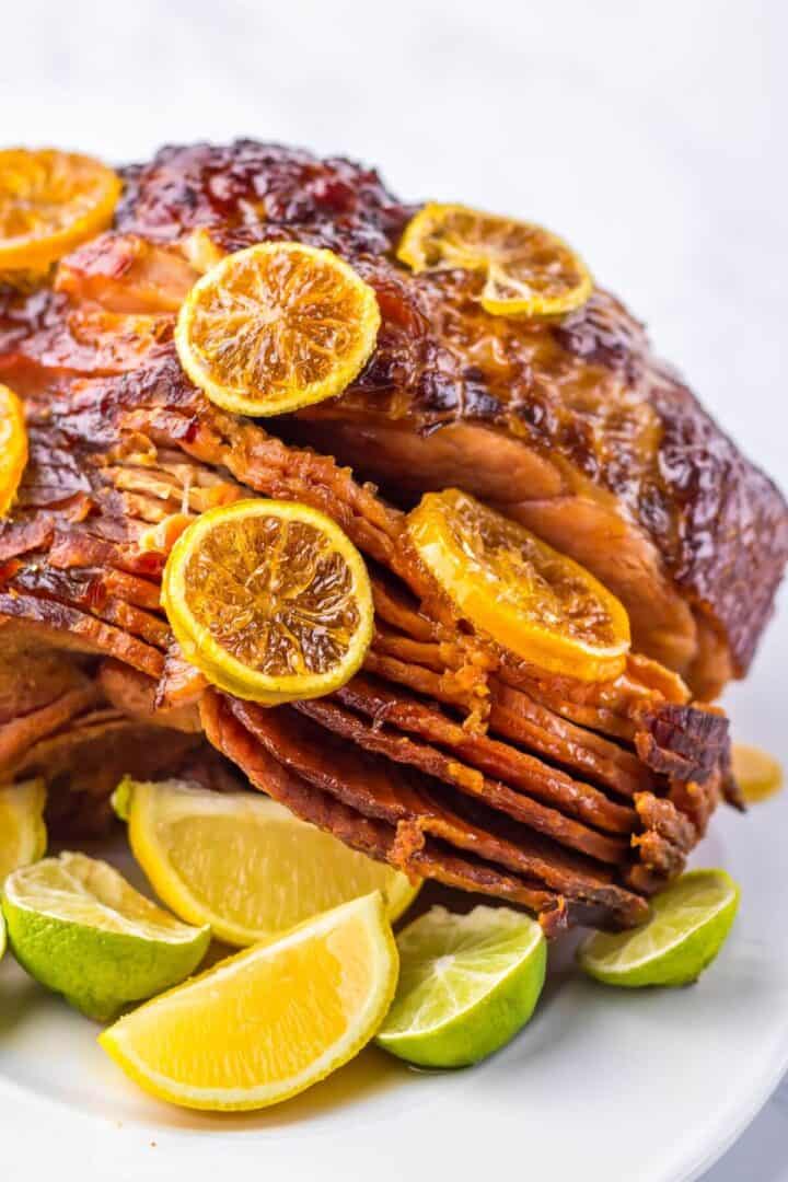 A Crock Pot Brown Sugar Pineapple Ham, adorned with orange slices and encircled by lime wedges, sits enticingly on a white plate.