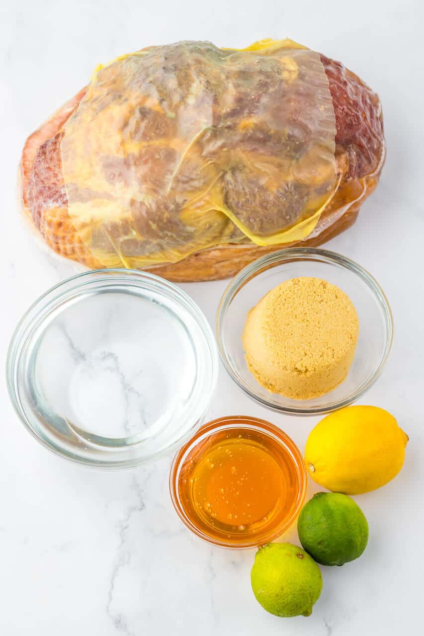 Crock Pot Brown Sugar Pineapple Ham sits elegantly beside bowls of water and brown sugar, accompanied by a jar of honey, and a lemon and lime, all beautifully arranged on a marble surface.