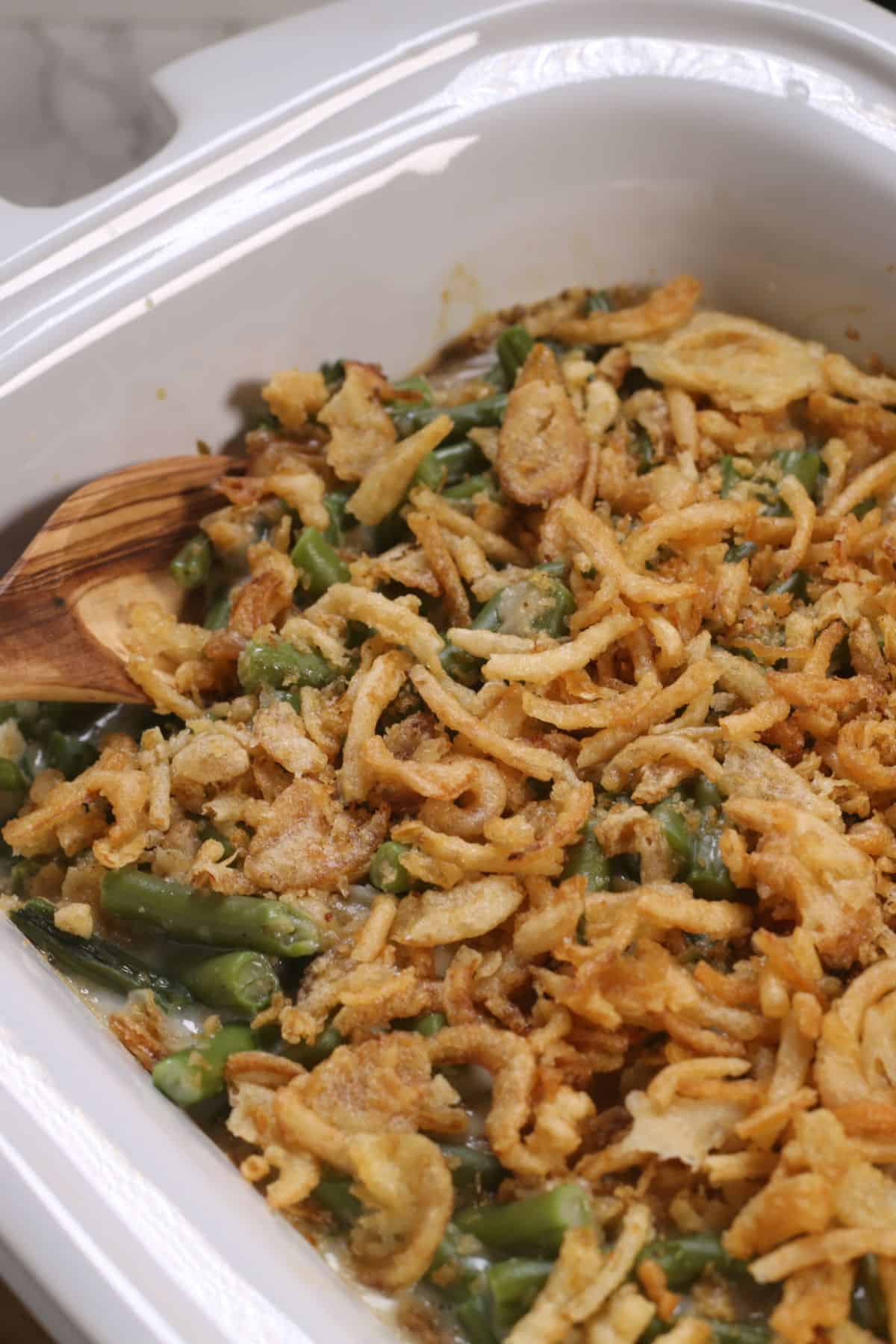 A Crock Pot Green Bean Casserole topped with crispy fried onions in a white baking dish, with a wooden spoon on the side.