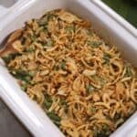 A white casserole dish filled with Crock Pot Green Bean Casserole, topped with crispy fried onions, sits beside a folded green cloth.