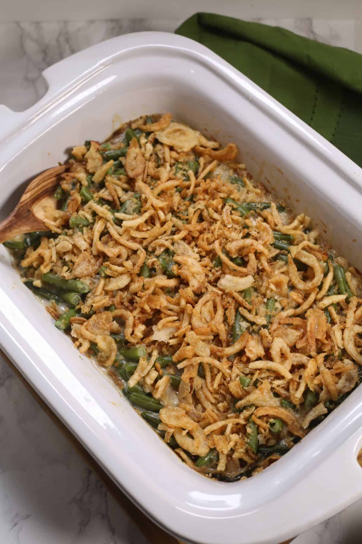 A white casserole dish filled with Crock Pot Green Bean Casserole, topped with crispy fried onions, sits beside a folded green cloth.